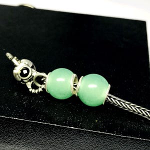 Könnte beinhalten: Ein silbernes Armband mit zwei hellgrünen, runden Perlen. Das Armband hat einen dekorativen Verschluss mit einem schwarzen Stein. Die Perlen sind durch silberne Ringe getrennt. Das Armband liegt auf einer schwarzen Oberfläche.