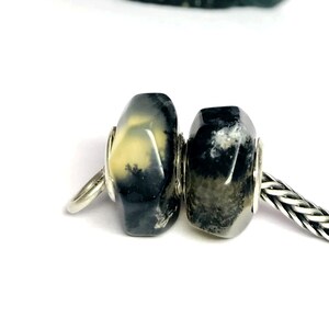 Könnte beinhalten: Zwei facettierte Perlen mit dendritischen Einschlüssen, die eine Mischung aus durchscheinenden cremefarbenen, grauen und schwarzen Mustern aufweisen. Die Perlen sind auf einer silberfarbenen Metallkette mit geflochtener Textur montiert.