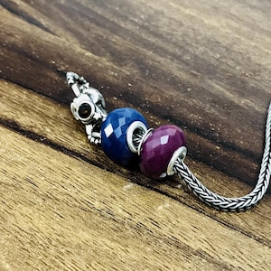 Könnte beinhalten: Ein silbernes Charm-Armband mit drei dekorativen Perlen. Die Perlen sind ein facettiertes Dunkelblau, ein facettiertes Dunkelviolett und ein silberner Charm mit einem dunkelbraunen Stein. Das Armband liegt auf einer Holzoberfläche.