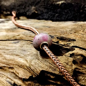 Könnte beinhalten: Ein kupferfarbenes Kettenarmband mit einer großen, runden, rosa-grauen Perle. Die Perle hat einen silberfarbenen Metallring um das Loch. Das Armband liegt auf einem Stück verwittertem Holz.