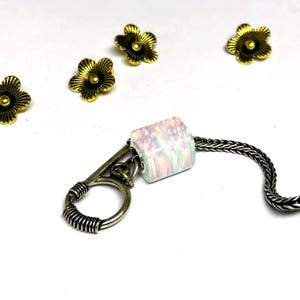 Könnte beinhalten: Ein Schlüsselanhänger mit einer weißen, schillernden, rechteckigen Perle, einem silberfarbenen Verschluss und einer geflochtenen Kette. Vier kleine, goldfarbene Blumenanhänger sind auf weißem Hintergrund um den Schlüsselanhänger herum verstreut.