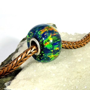 May include: A close-up of a bead with a vibrant, swirling pattern of blue, green, and orange. The bead is strung on a brown, braided leather cord. The bead has a silver-coloured metal ring.