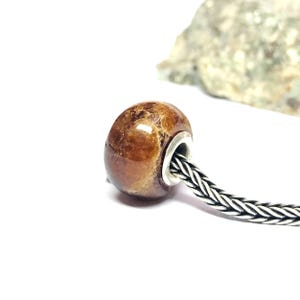 Könnte beinhalten: Eine runde, polierte braune Edelsteinperle mit glatter, gesprenkelter Oberflächenstruktur. Die Perle hat ein mittiges Loch mit einer silberfarbenen Metallöse und ist auf eine flexible, geflochtene silberfarbene Metallkette aufgezogen.