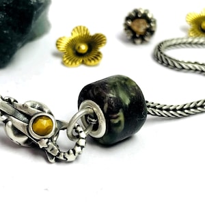 Könnte beinhalten: Nahaufnahme eines Armbands mit einer dunkelgrünen und schwarzen Perle. Das Armband hat einen silbernen Verschluss mit einem gelben Stein und eine silberne Kette. Goldene blütenförmige Perlen und ein silberner Ohrstecker befinden sich im Hintergrund.