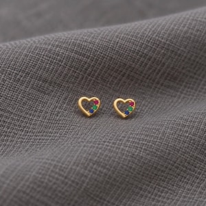 Könnte beinhalten: Ein Paar herzförmige Ohrstecker aus Gold. Jeder Ohrstecker hat eine hohle Mitte und ist mit drei kleinen, runden, mehrfarbigen Edelsteinen in Pink, Grün und Blau verziert, die in einer diagonalen Linie entlang einer Seite des Herzrahmens angeordnet sind.