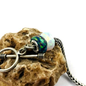 May include: A close-up of a silver charm bracelet with three opal beads. The beads are iridescent with colors of blue, green, and white. The bracelet is resting on a rough, tan stone.