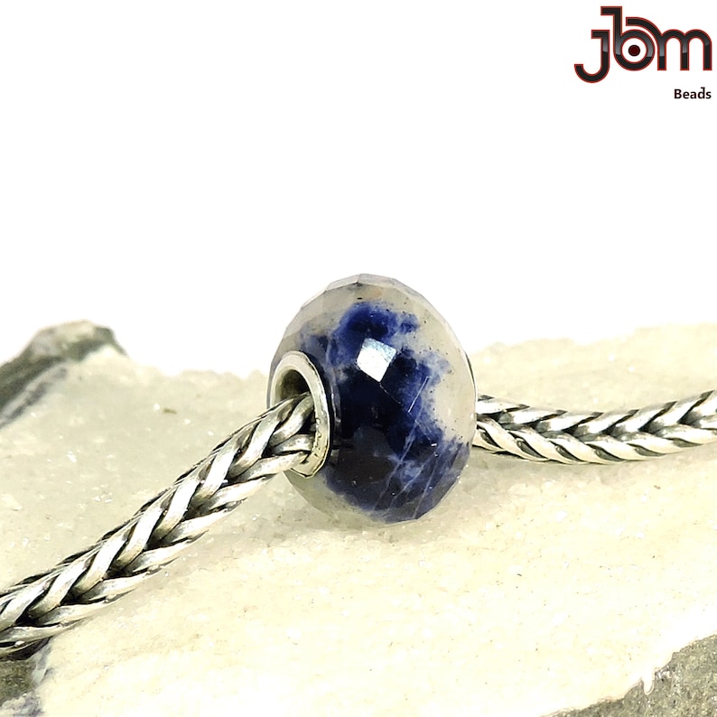 K&ouml;nnte beinhalten: Eine facettierte Perle mit blau-wei&szlig;em Muster, auf einer silbernen Kette aufgef&auml;delt. Die Perle hat einen Kern aus silberfarbenem Metall. Das Bild enth&auml;lt den Text "jbm Beads" in der oberen rechten Ecke.
