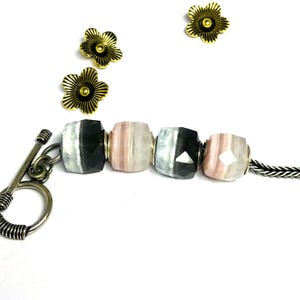 May include: A close-up of a bracelet featuring faceted beads in shades of black, gray, and pink. The bracelet has a silver toggle clasp and is adorned with three gold-colored flower charms.