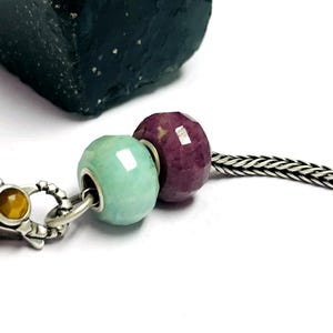 Könnte beinhalten: Nahaufnahme eines Armbands mit zwei facettierten Perlen: eine hellgrüne und eine tiefrote. Die Perlen sind an einer Silberkette aufgereiht, mit einem dekorativen Verschluss mit einem gelben Stein. Im Hintergrund befindet sich ein dunkler, strukturierter Stein.