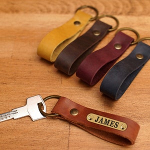 Peut inclure: Cinq porte-clés en cuir avec des anneaux en laiton et une clé attachée à l'un des porte-clés. Les porte-clés sont de différentes couleurs : jaune, marron, bordeaux, bleu foncé et marron. Le porte-clés marron a une étiquette en laiton avec le nom "JAMES" gravé dessus.