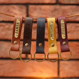 Puede incluir: Cinco llaveros de cuero de varios colores, cada uno con una placa de latón. Los llaveros son marrones, marrón oscuro, negro, amarillo y burdeos. Las placas están grabadas con nombres, iniciales y coordenadas. El texto "DRIVE SAFE" también es visible.