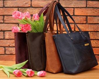 May include: Four leather tote bags in different colors: burgundy, dark brown, brown, and navy blue. The bags are arranged in a row with the burgundy bag on the left and the navy blue bag on the right. A bouquet of pink tulips is in front of the bags.