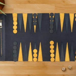 May include: A navy blue backgammon board with yellow and grey detailing, featuring a zip closure and a roll-up design. Includes black and yellow game pieces and two dice. The board is displayed on a wooden surface.