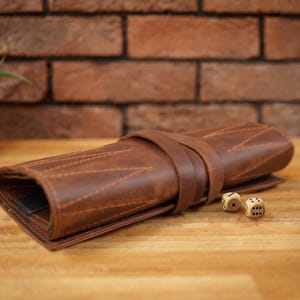 Könnte beinhalten: Eine aufgerollte braune Würfelbox aus Leder mit einem Riemen, die auf einer Holzoberfläche liegt. Zwei Würfel liegen neben der Box. Der Hintergrund zeigt eine Backsteinmauer und eine kleine Topfpflanze.