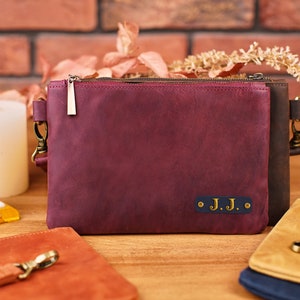 May include: A burgundy leather clutch with a zipper closure and a gold metal pull. The clutch has a dark blue leather patch with the initials "J.J." in gold lettering.