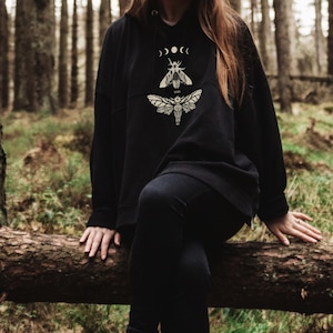 Op de afbeelding: Zwarte hoodie met een grafische print van een nachtvlinder en een kever met maanfasen en de tekst "Forest Wings".