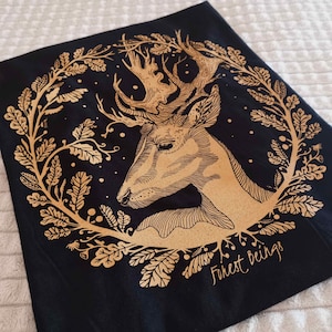 Könnte beinhalten: Ein schwarzes T-Shirt mit einem goldenen Foliendruck eines Hirschkopfes, umgeben von Blättern und dem Text "Forest beings".