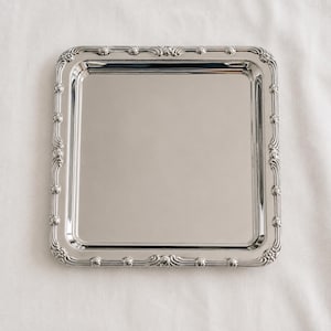Peut inclure: Un plateau de service carré de couleur argentée avec une surface polie et réfléchissante. Le plateau présente un bord surélevé orné d'un motif répétitif de petites sphères et de lignes courbes et fluides sur les quatre côtés du cadre.