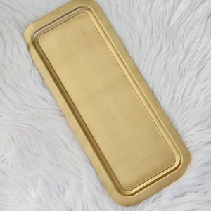 Puede incluir: Una bandeja rectangular de metal dorado con un borde ligeramente elevado. La bandeja tiene esquinas redondeadas y un acabado cepillado. La superficie es lisa y refleja la luz. La bandeja está diseñada para servir o exhibir.