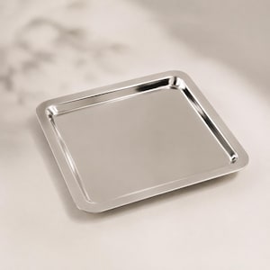 Puede incluir: Una bandeja cuadrada de acero inoxidable pulido con una superficie lisa y reflectante y esquinas ligeramente redondeadas. La bandeja presenta una base plana y un borde estrecho elevado que enmarca el área central, creando un diseño metálico.