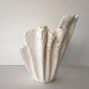 May include: A white, textured ceramic vase with an unusual, organic shape. The vase has a wide, fluted design with a rough, uneven surface. The overall form is reminiscent of a seashell or draped fabric, suitable for home decor.
