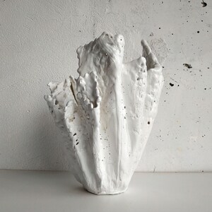 May include: A white ceramic vase with a textured, abstract design. The vase has a rough, uneven surface with multiple openings and a flared shape. The sculpture is set against a white background, highlighting its unique form.