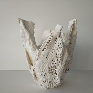 May include: A white ceramic vase with a textured, lace-like pattern. The vase has an organic, open shape with irregular edges, creating a unique, handcrafted appearance. The color is a soft, off-white, suitable for home decor.