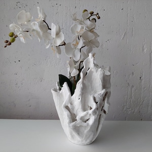 May include: A white, textured vase with an organic, open design, holding white orchid flowers with green accents. The vase sits on a white surface against a textured white wall, creating a minimalist aesthetic.