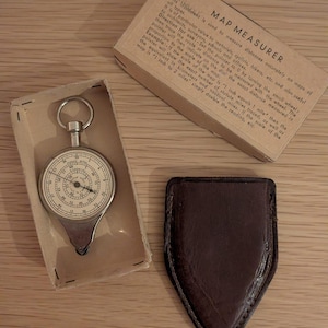 Könnte beinhalten: Ein Vintage-Kartenmessgerät mit rundem Zifferblatt, untergebracht in einer Pappschachtel, neben einem braunen Lederetui. Das Zifferblatt hat Zahlen und Markierungen. Auf der Schachtel steht "MAP MEASURER".