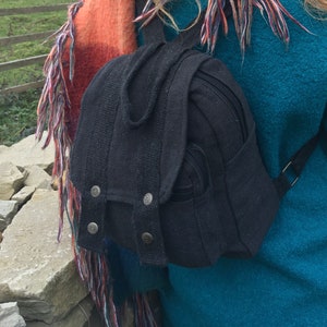 May include: A black backpack with a flap closure secured by three silver buttons. The small bag has a zippered main compartment and a small side pocket. The backpack is made of a textured fabric and has adjustable straps.