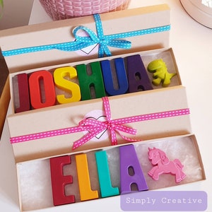 May include: Two gift boxes with rainbow crayon letters spelling out the names "Joshua" and "Ella". Each box is tied with a ribbon and includes a small toy, a green dinosaur and a pink unicorn.
