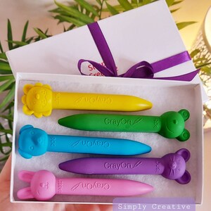 May include: A set of five colorful crayons shaped like animals. The crayons are in a white box with a purple ribbon. The crayons are labeled with the words "Crayon" and the name of the animal. The animals are a lion, a dog, a rabbit, a frog, and a mouse.
