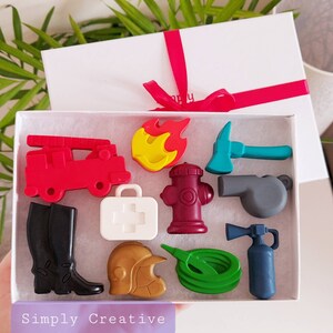 May include: A white gift box with a red ribbon contains colorful wax crayons shaped like firefighting equipment. The set includes a red fire truck, a yellow and red flame, a teal axe, a white first aid kit, and a maroon fire hydrant.