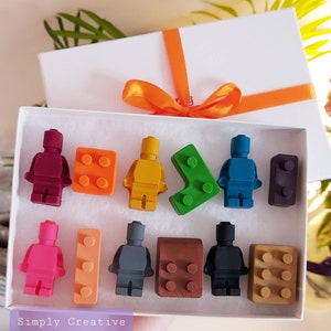 May include: A white gift box tied with an orange ribbon, containing colorful crayon figures shaped like toy bricks and people. The crayons are in various colors, including red, orange, yellow, green, blue, pink, gray, brown, and black. The text "Simply Creative" is visible.