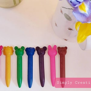 May include: Eight colorful crayons shaped like animals. The crayons are pink, yellow, green, blue, purple, red, brown, and orange. The crayons are arranged in a row on a white surface. The text "Simply Creative" is visible in the bottom right corner of the image.