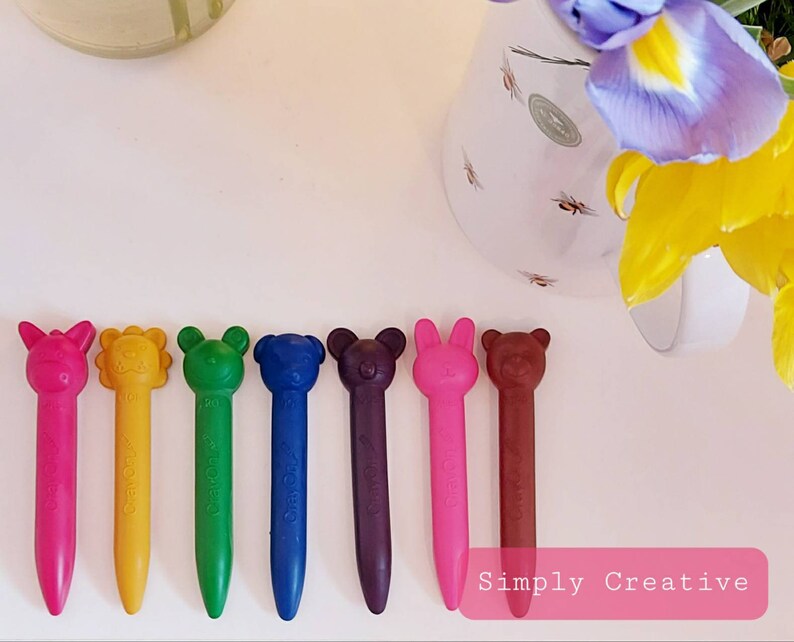 May include: Eight colorful crayons shaped like animals. The crayons are pink, yellow, green, blue, purple, red, brown, and orange. The crayons are arranged in a row on a white surface. The text "Simply Creative" is visible in the bottom right corner of the image.