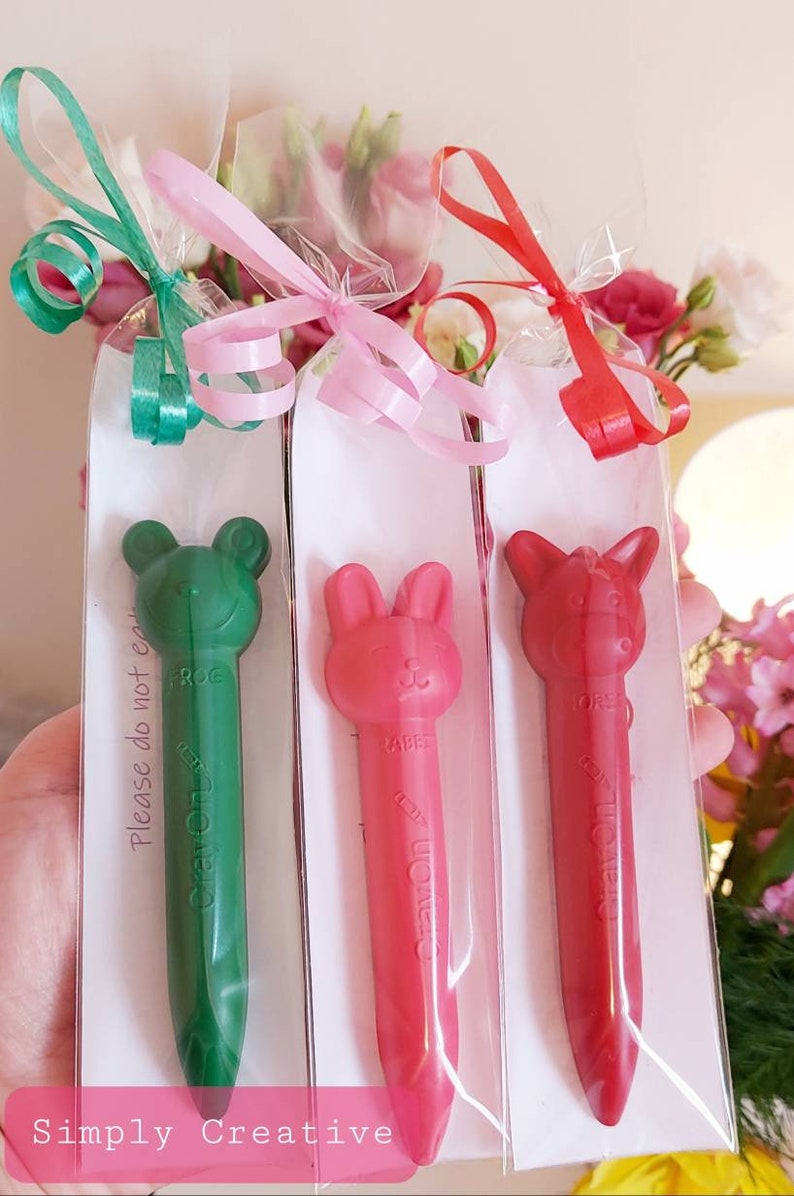 May include: Three individually wrapped crayons with animal faces. The crayons are green, red, and pink. The green crayon is a frog, the red crayon is a rabbit, and the pink crayon is a horse. Each crayon has the word "Crayon" written on it. The packaging has a white background with the text "Please do not eat" written on it. The packaging is tied with a ribbon.