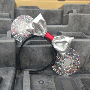 Pode incluir: Orelhas de Minnie Mouse com glitter prateado, laço prateado e fita vermelha. As orelhas são decoradas com glitter colorido.