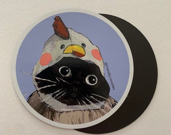 May include: A black and white cat wearing a white and yellow chicken costume with pink cheeks. The cat is looking at the camera. The background is a light purple color. The text "@friendly_noodles" is in the bottom right corner of the image.