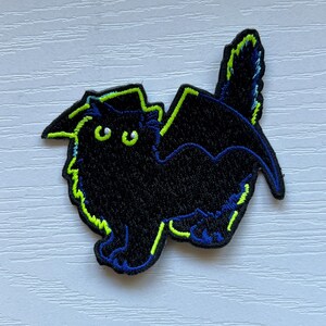 May include: Black embroidered iron-on patch of a cat with bat wings outlined in green and blue.