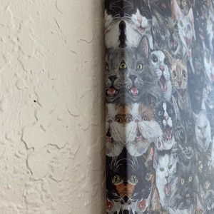 May include: A canvas print featuring a collage of cat faces with various expressions. The cats are depicted in a range of colors, including gray, black, white, and brown. The artwork is mounted on a wooden frame and displayed against a textured white wall.