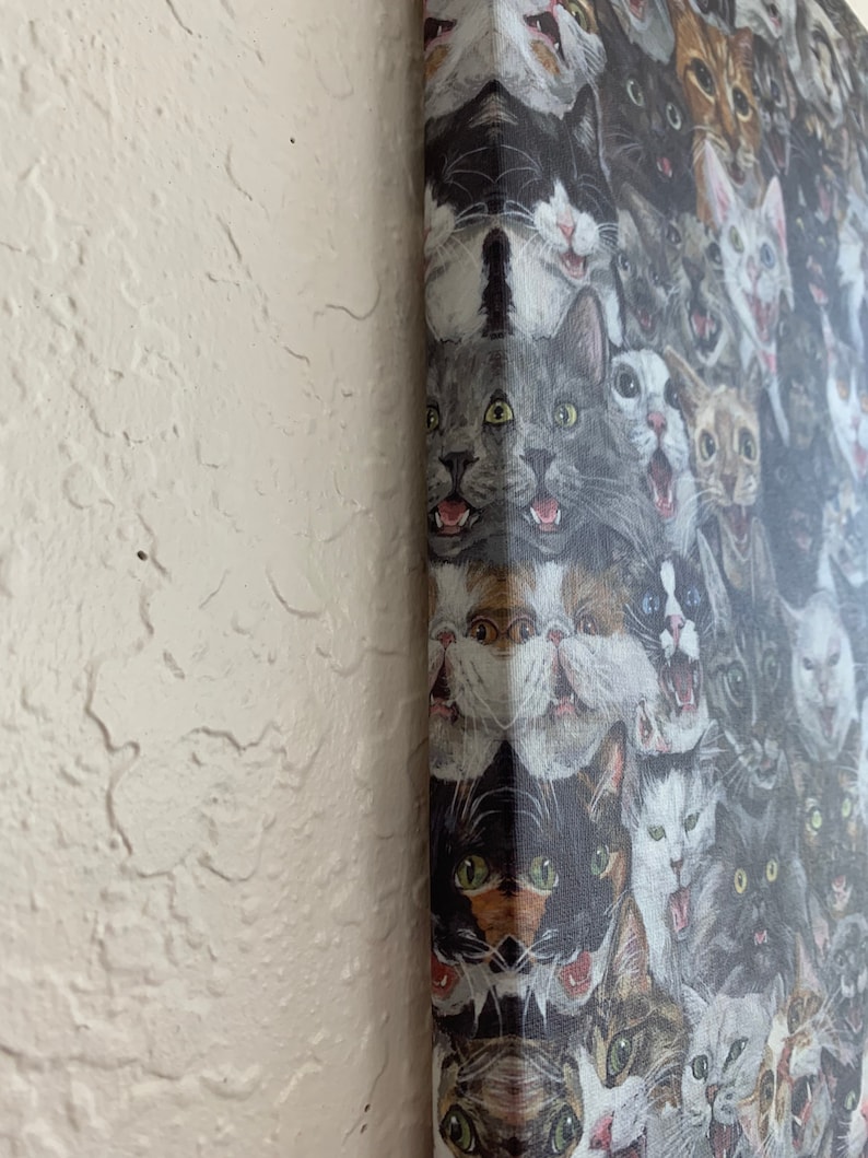 May include: A canvas print featuring a collage of cat faces with various expressions. The cats are depicted in a range of colors, including gray, black, white, and brown. The artwork is mounted on a wooden frame and displayed against a textured white wall.