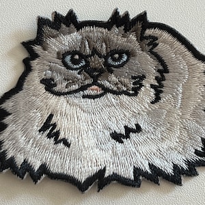 Peut inclure: Un patch brodé représentant un chat duveteux aux yeux noirs et au nez rose, dans les tons gris et blanc.
