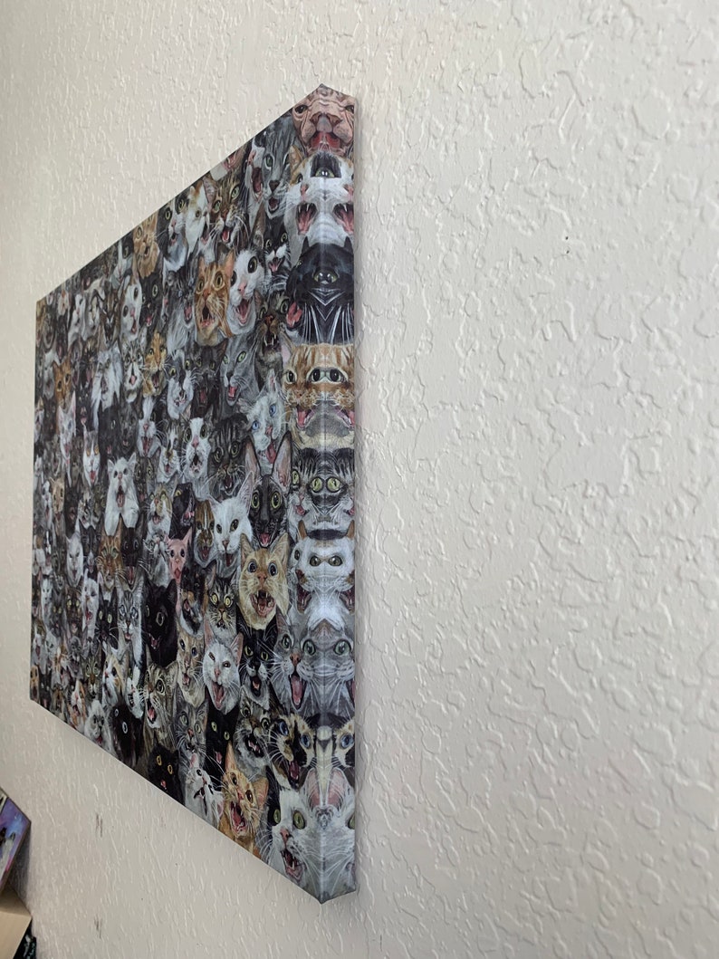 May include: A canvas print featuring a collage of numerous cat faces, each with a unique expression. The cats' fur colors vary, including black, white, gray, and tabby patterns. The artwork is mounted on a wooden frame and displayed on a white wall.