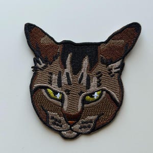 May include: Embroidered patch featuring a cat's face. The patch has a brown and tan color scheme with black accents. The cat's eyes are yellow with white highlights, and the nose is a light brown color. The patch is designed to be sewn or ironed onto fabric.