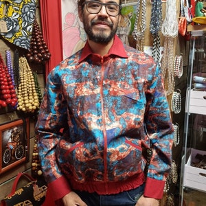 May include: A man wearing a blue and red patterned jacket with a zipper closure. The jacket has a red collar and red cuffs. The man is wearing blue jeans and has a beard and glasses.