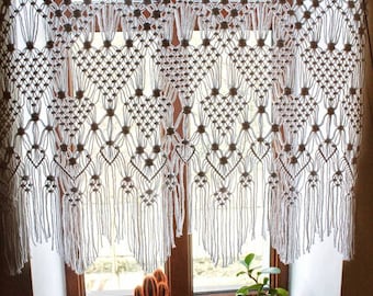 May include: A white macrame curtain hangs in a window with a wooden frame. The curtain has a geometric pattern and is made of knotted cords. Two potted plants sit on the windowsill, one with a cactus and the other with green leaves.