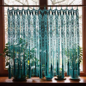 May include: Teal macrame curtain hanging in front of a window. The curtain is made of knotted cords, with a detailed pattern at the top and long, flowing strands. The window is framed by a wooden frame, with plants visible in the background.