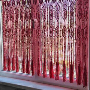 May include: A pink macrame curtain with tassels hangs in a window. The curtain is made of a series of knotted cords, creating a geometric pattern. The tassels are long and flowing, adding a touch of elegance to the curtain.