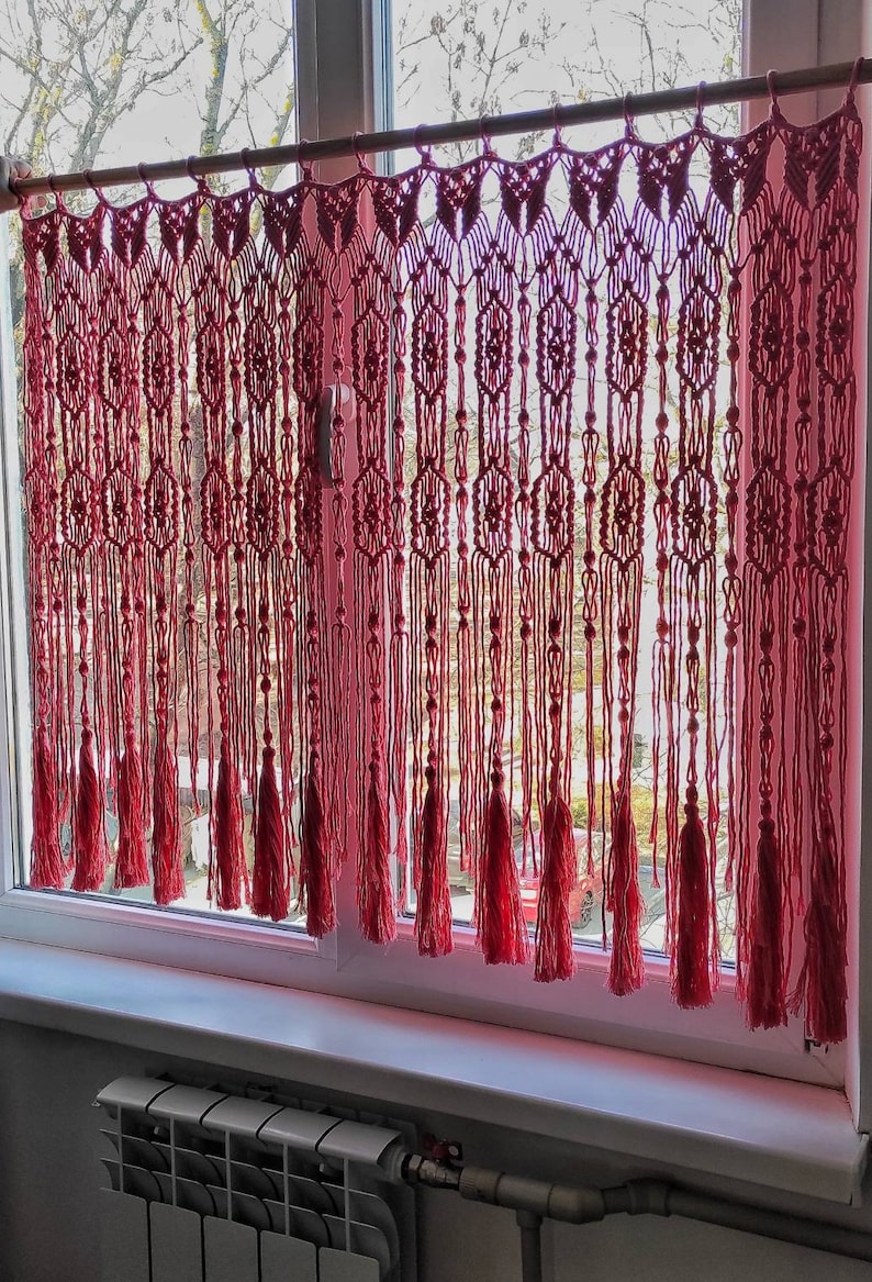 May include: A pink macrame curtain with tassels hangs in a window. The curtain is made of a series of knotted cords, creating a geometric pattern. The tassels are long and flowing, adding a touch of elegance to the curtain.